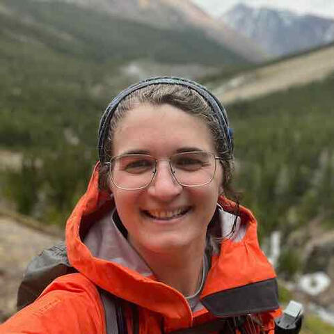 Profile picture of Kira McLean wearing orange rain jacket with blurred mountain landscape behind her.